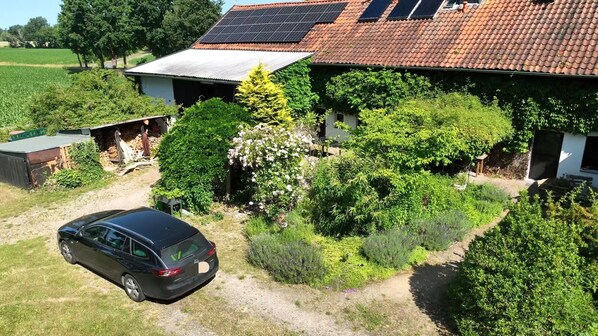 Una casa con paneles solares, un coche aparcado delante y un jardín con diversas plantas y árboles.