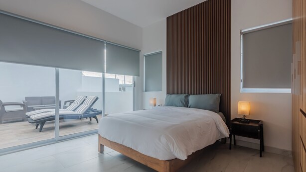 A modern bedroom with a large bed, a small bedside table, a window, and a view of a balcony with seating.