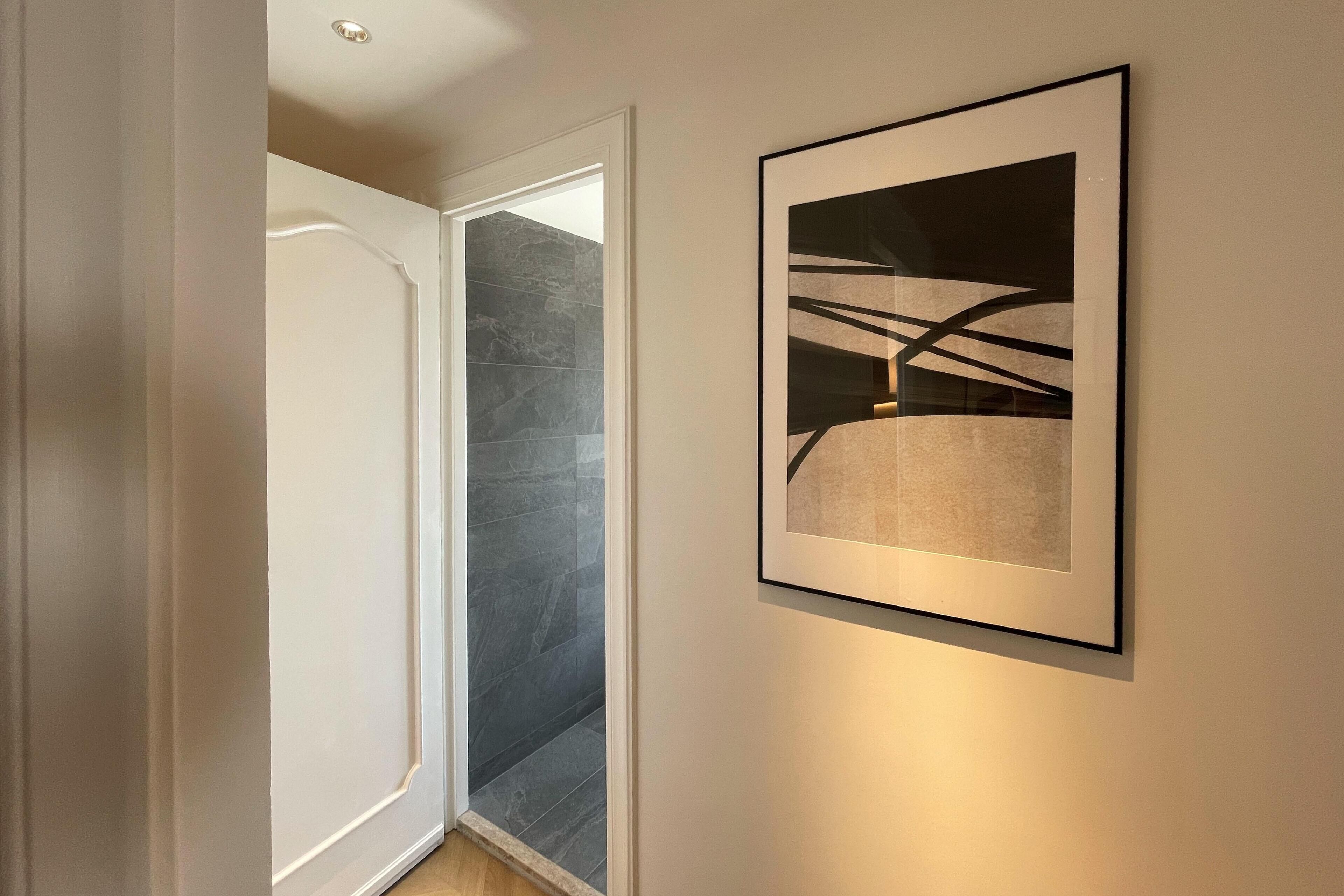 A hallway with a door leading to a room with dark grey tiles, a framed abstract artwork on the wall, and a light fixture above.