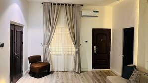 A modern living room with a wooden floor, a brown leather sofa, a beige armchair, and a large window with beige curtains. - 2bedroom luxury apt 24hrs light/wifi in lekki (Lekki)