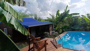 A wooden deck with a pool, surrounded by lush greenery and a blue-roofed structure.