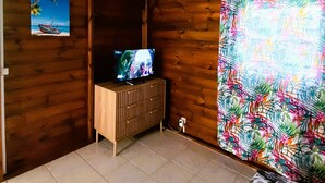 A wooden cabin room with a television on a wooden cabinet, a beach painting on the wall, and a tropical patterned curtain. - belle créole (Anse-Bertrand)