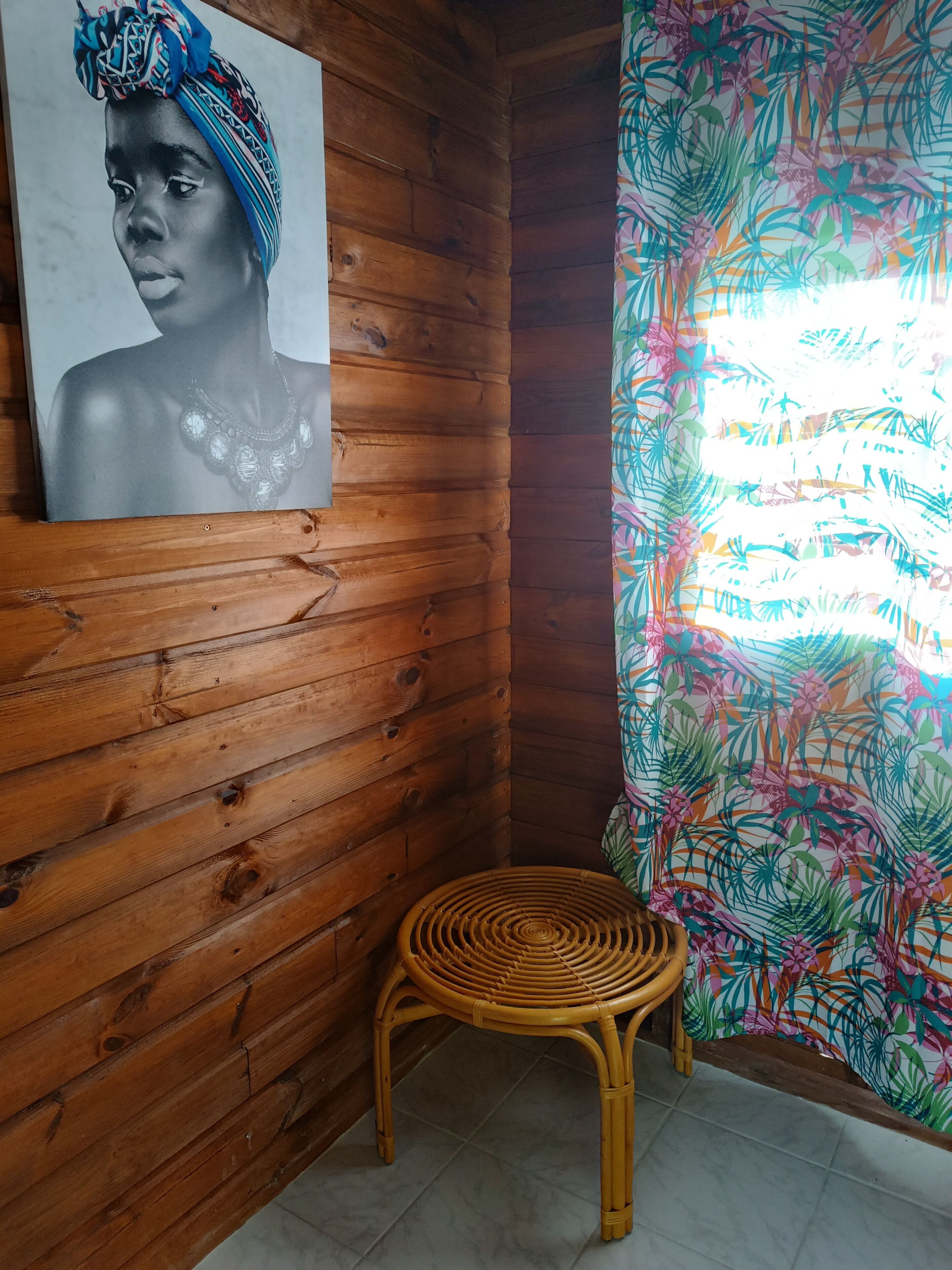 En stuga med träpanel på insidan, en affisch på väggen, en flätad stol och ett fönster med en gardin med tropiskt mönster.
