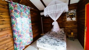 A wooden cabin room with a bed, a mosquito net, a colorful curtain, and a small table with a lamp. - belle créole (Anse-Bertrand)