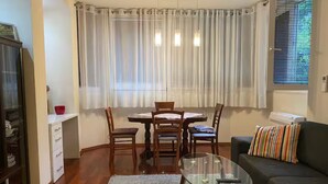 Un salon avec une table basse en verre, un canapé agrémenté d’un coussin à rayures, une table à manger avec des chaises, et une fenêtre avec des rideaux.