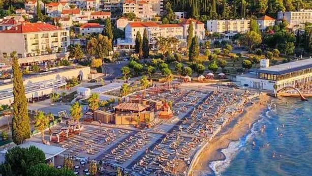 Une vue aérienne d’une ville côtière avec une plage où l’on aperçoit des rangées de chaises longues et de parasols, le tout entouré de bâtiments résidentiels et surplombé par un pont.
