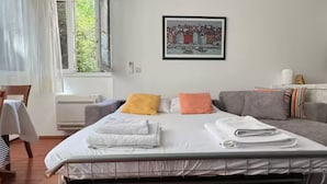 Une chambre d’hôtel avec un lit, un canapé gris, un tableau encadré au mur et une fenêtre avec des rideaux.