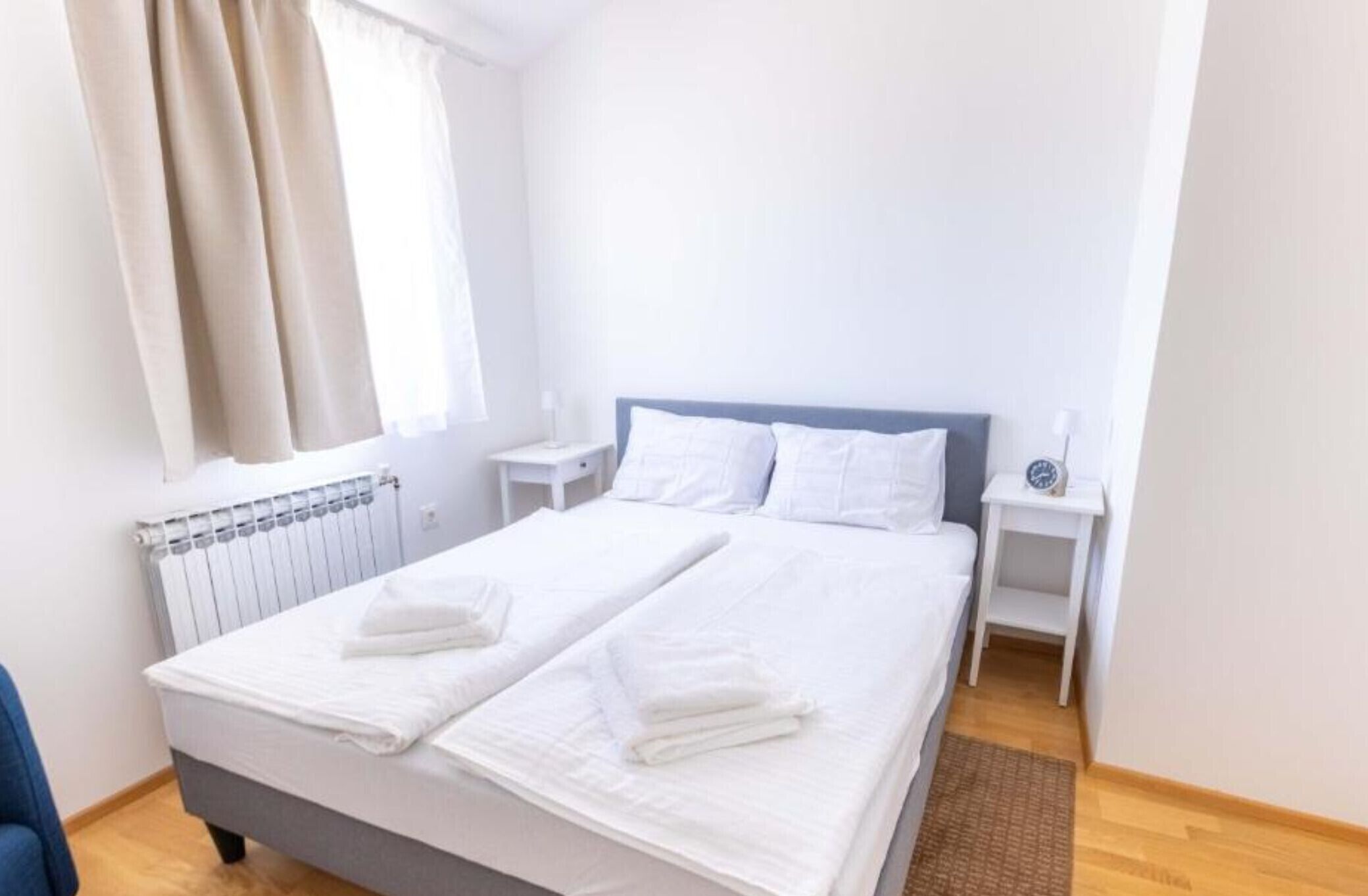 Ein ordentlich bezogenes Bett mit weißen Leinen und Handtüchern, ein Nachttisch mit einer Uhr und ein Fenster mit Vorhängen.