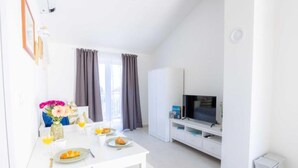Ein heller, moderner Wohnraum mit einem weißen Sofa, einem Fernseher auf einem Schrank und einer Essecke, ein Tisch ist für das Frühstück gedeckt.