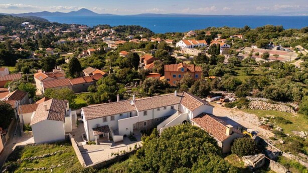 Eine Vogelperspektive auf eine Küstenstadt mit terrassenförmig angelegten Häusern, einem markanten weißen Gebäude mit rotem Dach sowie freiem Blick auf das Meer und die dahinterliegenden Berge.