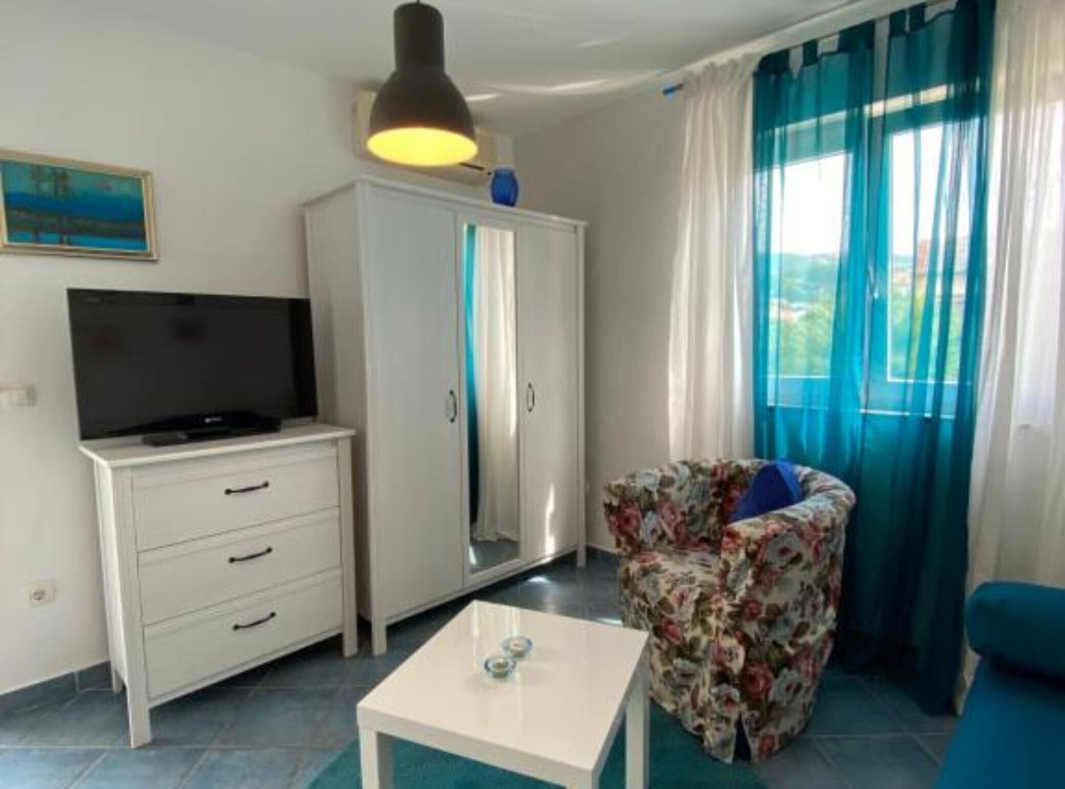 A room with a TV on a white cabinet, a floral chair, a white table, and a wardrobe.