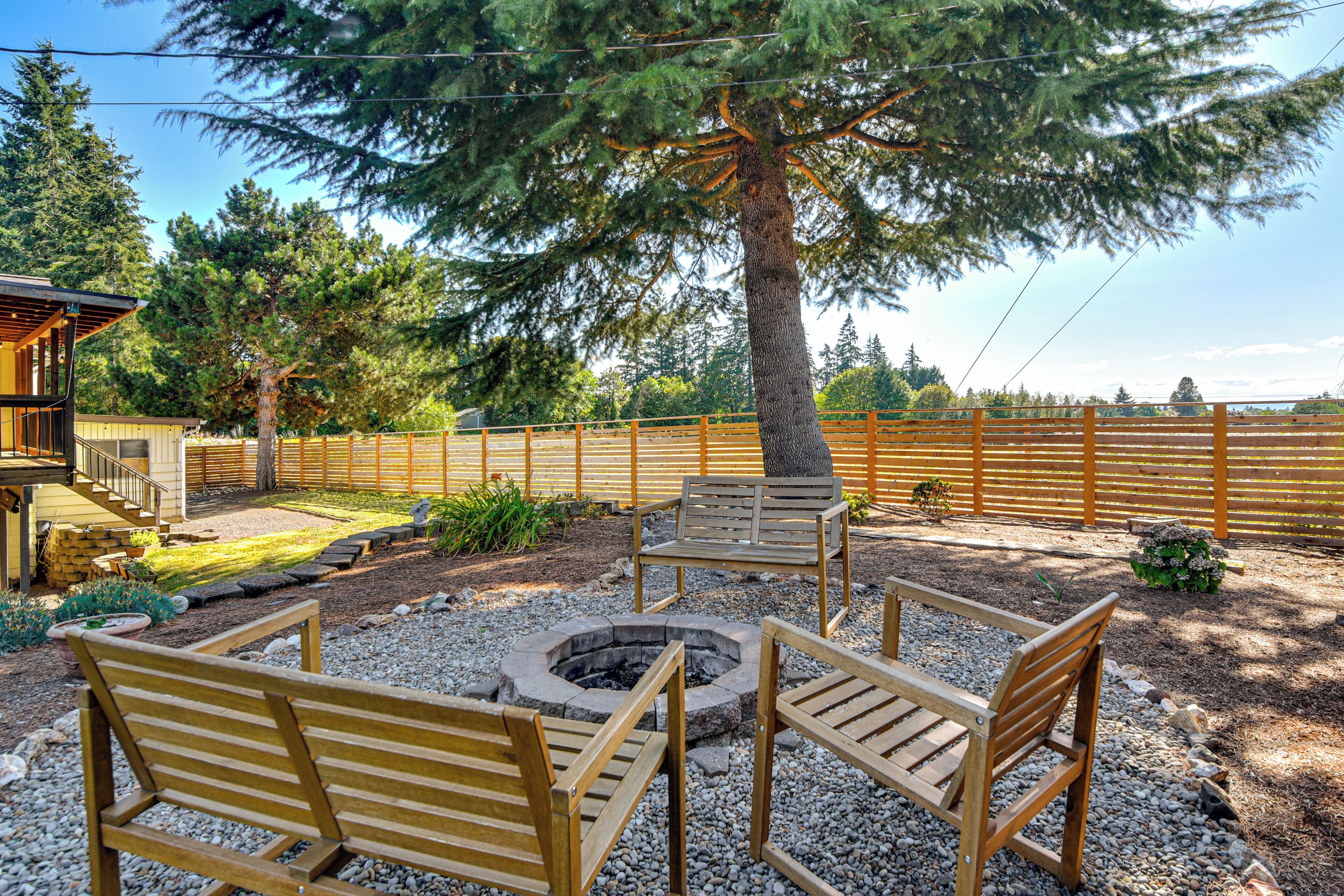 An outdoor seating area with wooden benches and a fire pit, surrounded by gravel and a wooden fence.