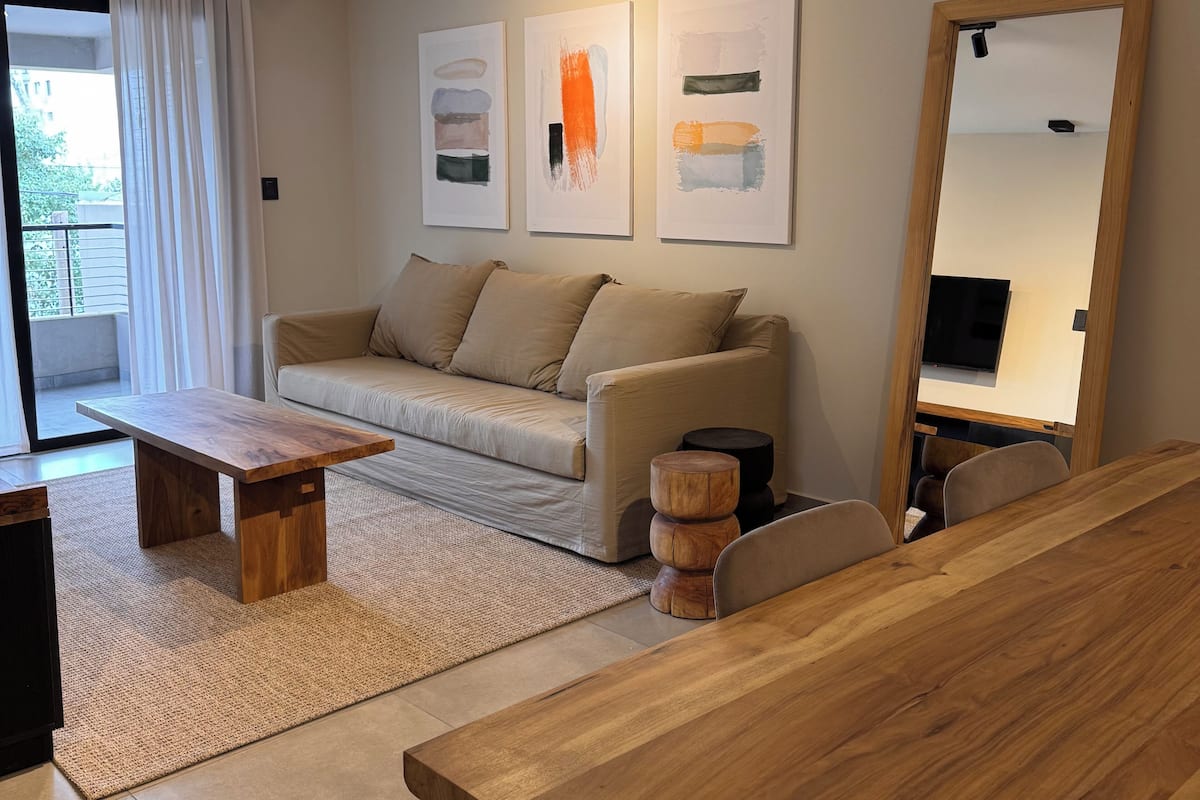 A modern living room with a sofa, coffee table, and wooden side table. There are abstract paintings on the wall and a large mirror.