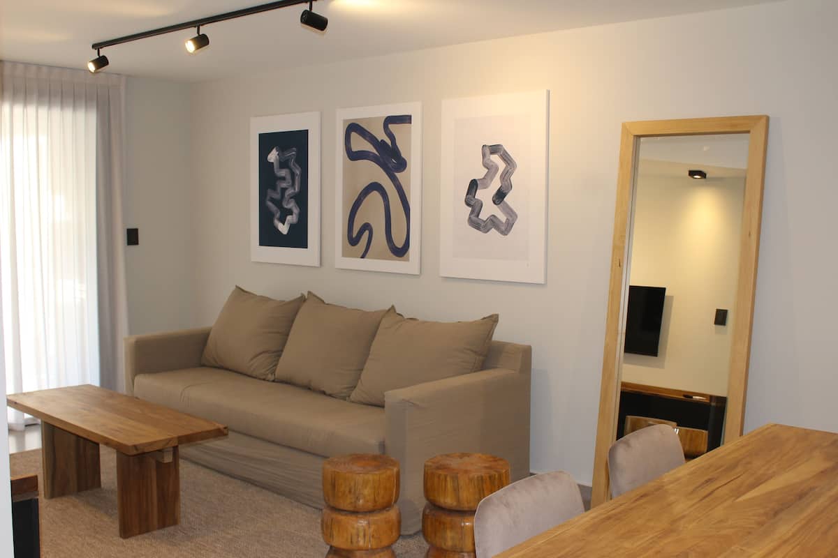 A modern living room with a sofa, wooden coffee table, and a dining table. There are three abstract art pieces on the wall and a mirror on the wall.
