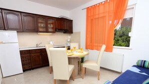 A compact kitchen with wooden cabinets, a dining table with chairs, and a window with orange curtains. - One-Bedroom Apartment With Terrace (A2) (Trogir)