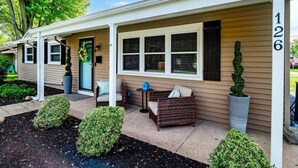 A single-story house with a porch, number 126, featuring a small seating area with a wicker chair and a table, surrounded by potted plants and a well-maintained garden. - Lake Cottage Retreat-Hot Tub-Game Room (Avon Lake)