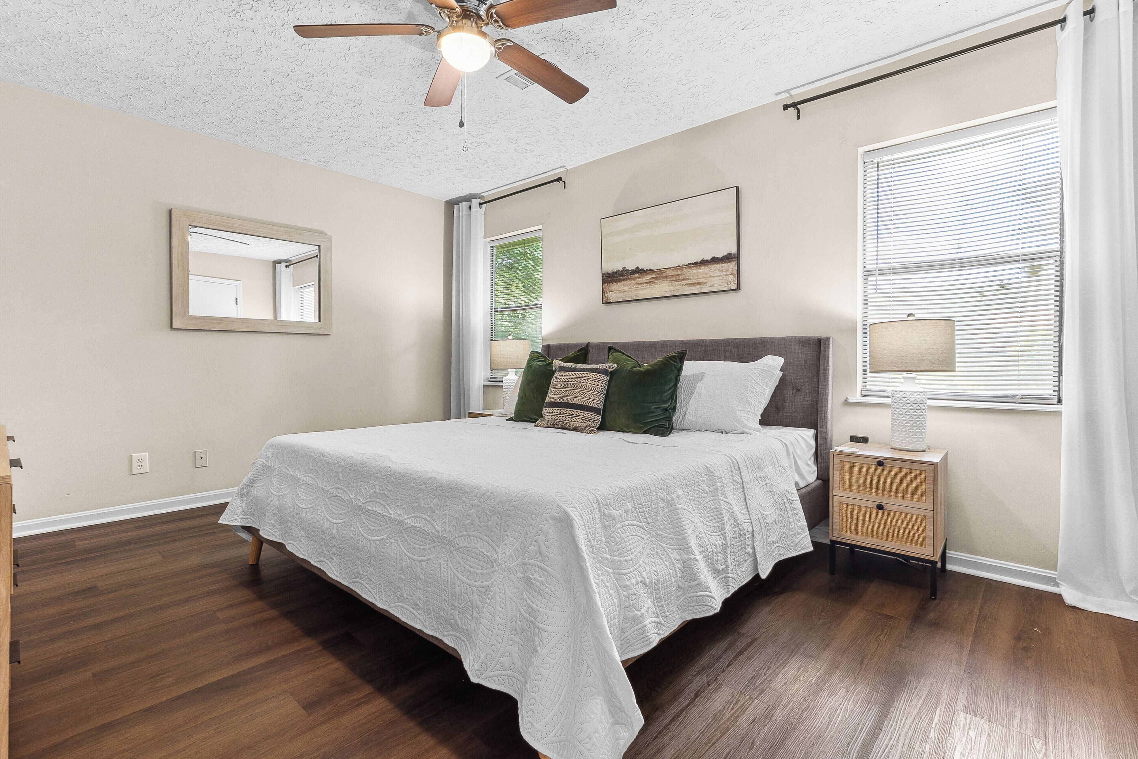A bedroom with a bed, a nightstand, a ceiling fan, a window with blinds, and a wall-mounted mirror.