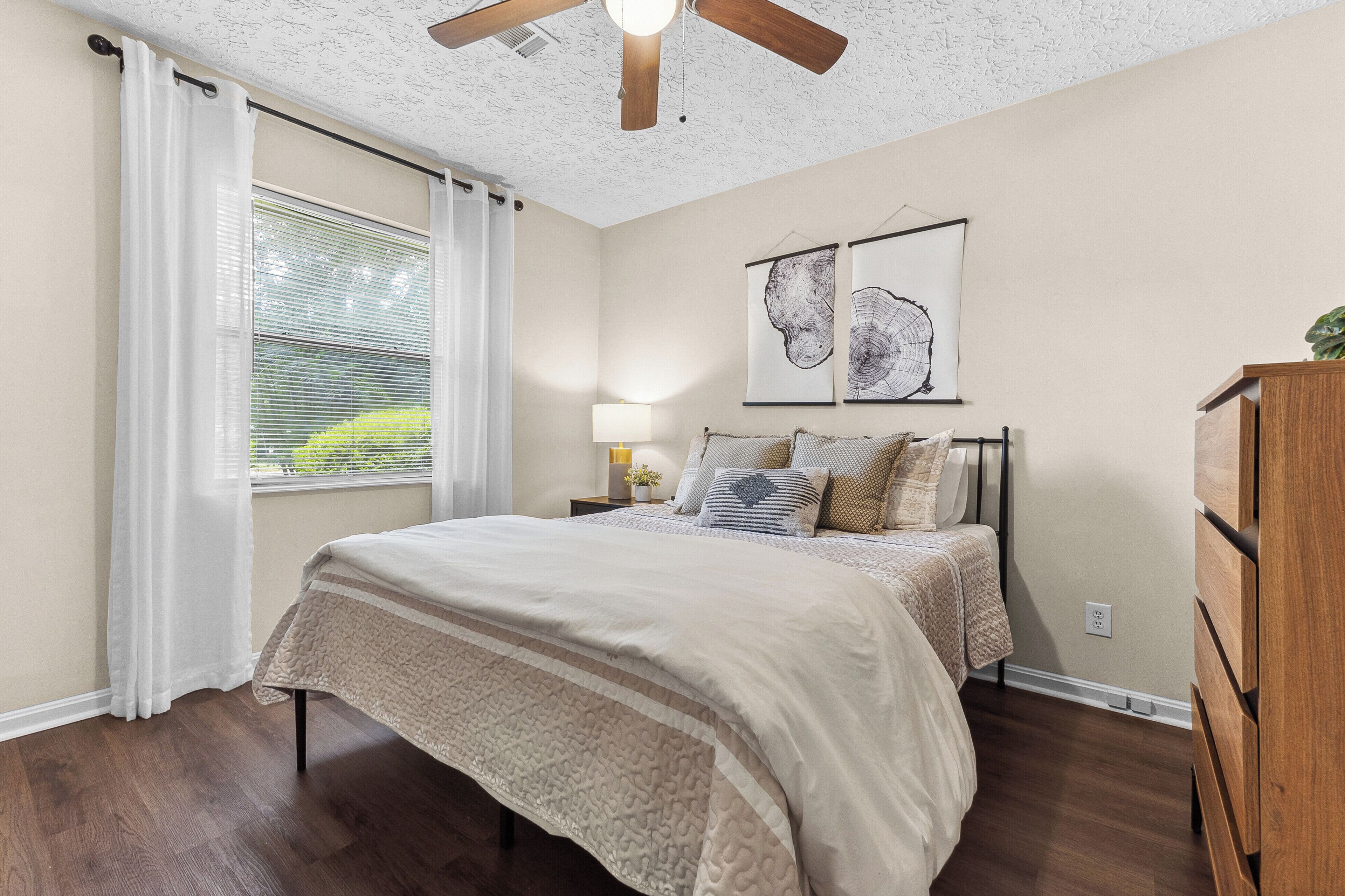 A bedroom with a bed, a ceiling fan, a window with curtains, and two wall-mounted artworks.