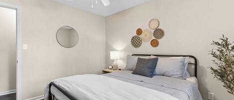 A bedroom with a bed, a ceiling fan, a bedside table with a lamp, and decorative wall art.