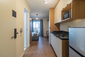 Corredor estreito que dá para um quarto com cama, uma pequena mesa e uma cadeira. Uma área de cozinha, com fogão, micro-ondas e geladeira, é visível à direita.