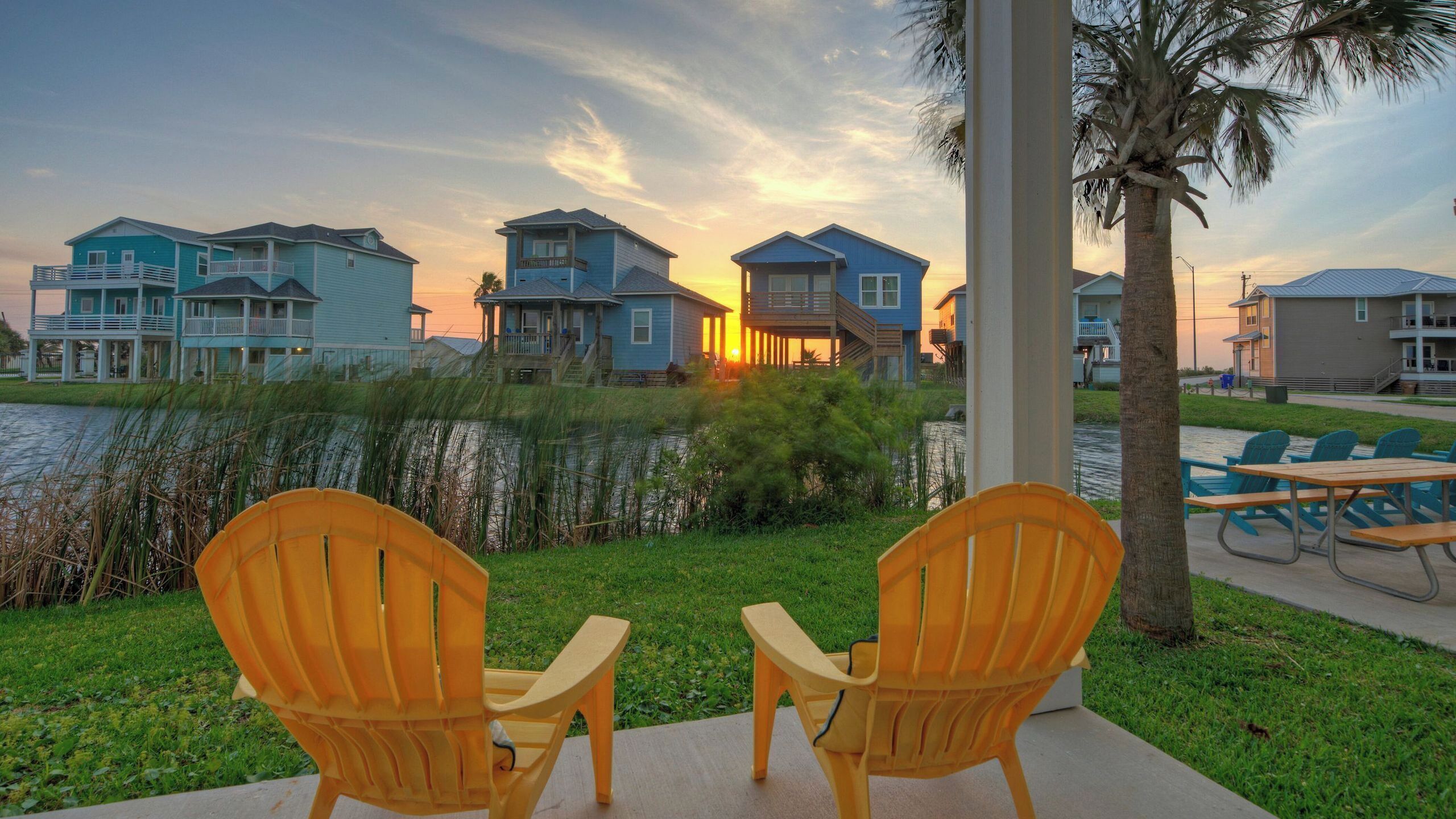 A waterfront property with multiple blue buildings, a grassy area, and two wooden chairs.