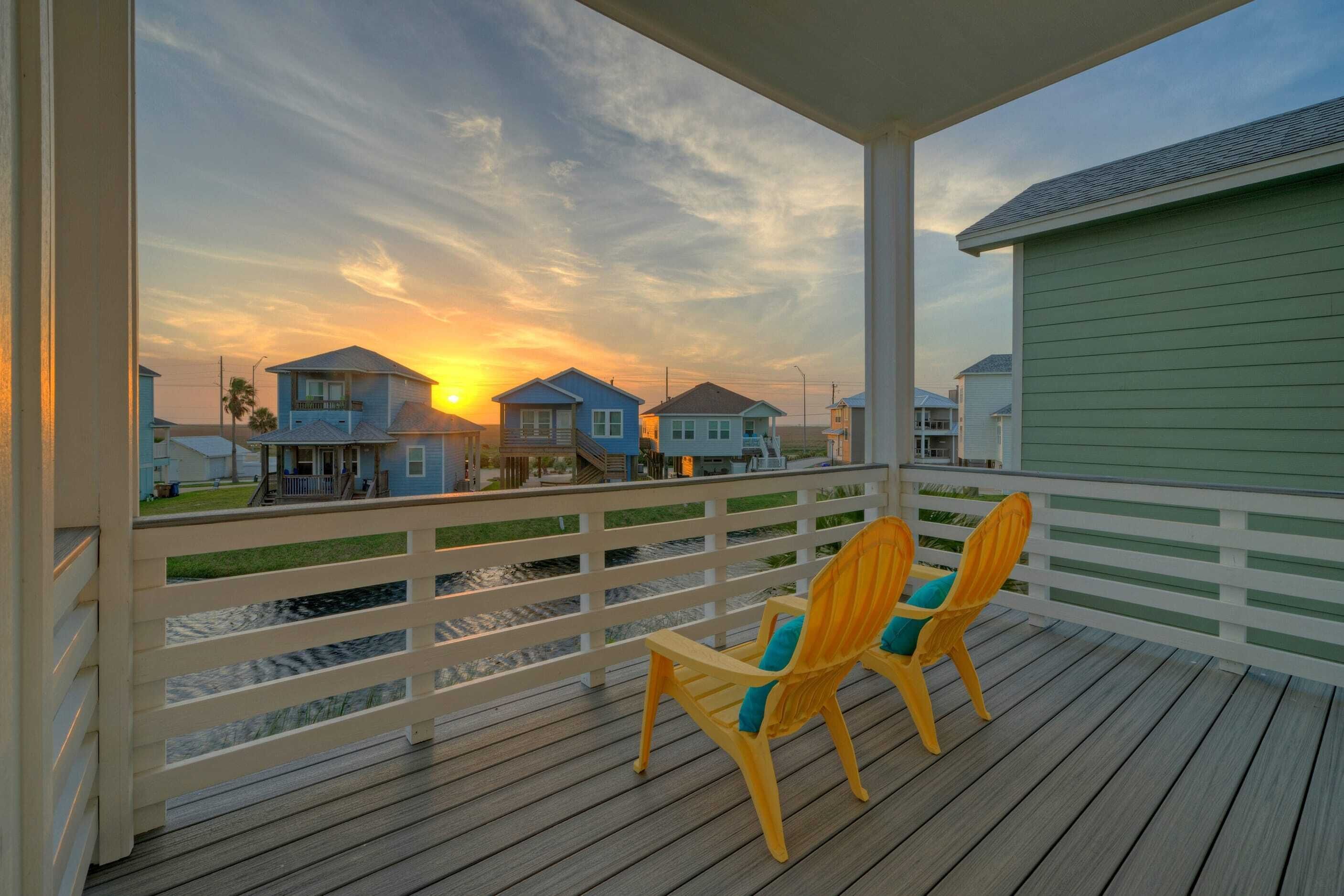 A wooden deck with two chairs and a table, overlooking a body of water and houses with a sunset in the background.
