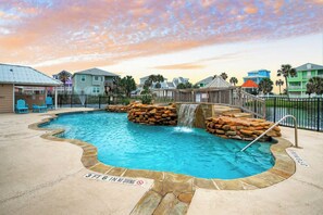 A swimming pool with a waterfall feature, surrounded by a wooden fence and a stone border.