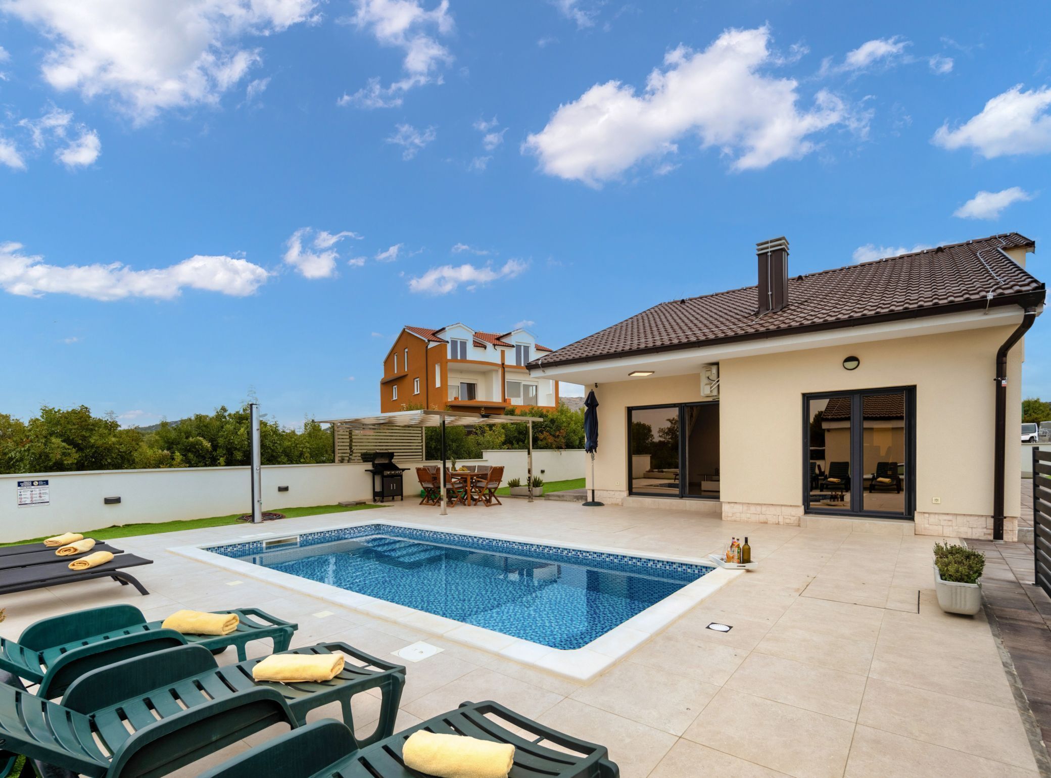 A swimming pool with lounge chairs, a covered patio, and a two-story house with a tiled roof.