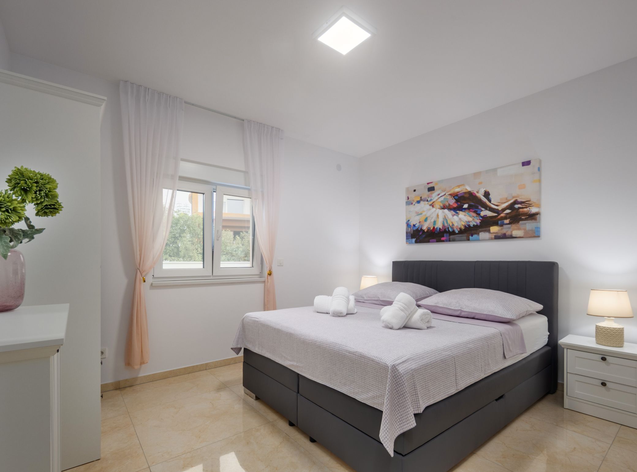 A modern bedroom with a bed, bedside tables, a window with curtains, a painting on the wall, and a potted plant.