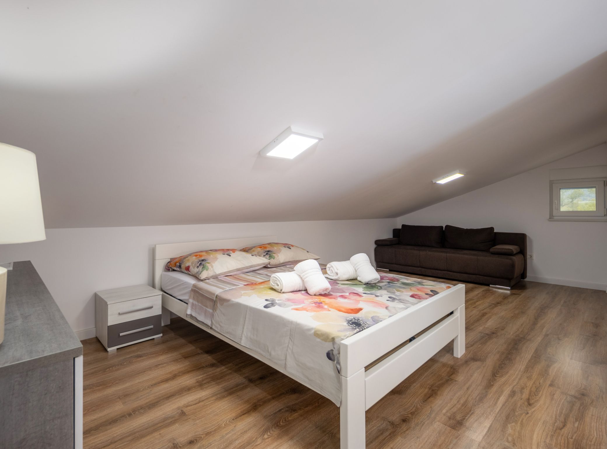 A bedroom with a bed, a nightstand, a dresser, a brown sofa, and a window.