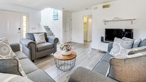 A modern living room with grey sofas, a coffee table with a floral centerpiece, a flat-screen TV, and a staircase leading to another room.