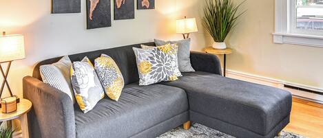 A modern living room with a grey sectional sofa, multiple patterned cushions, a world map artwork, a potted plant, and a window with a view of the street.