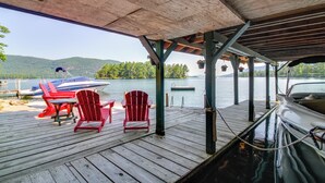 Un muelle de madera con sillas Adirondack rojas, un bote y vistas a un lago y montañas.