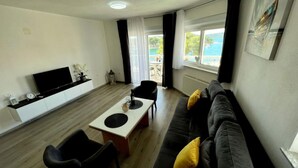 A modern living room with a flat-screen TV, a sofa with yellow cushions, a dining table, and a view of the sea through large windows. - Villa Tomi (Trogir)