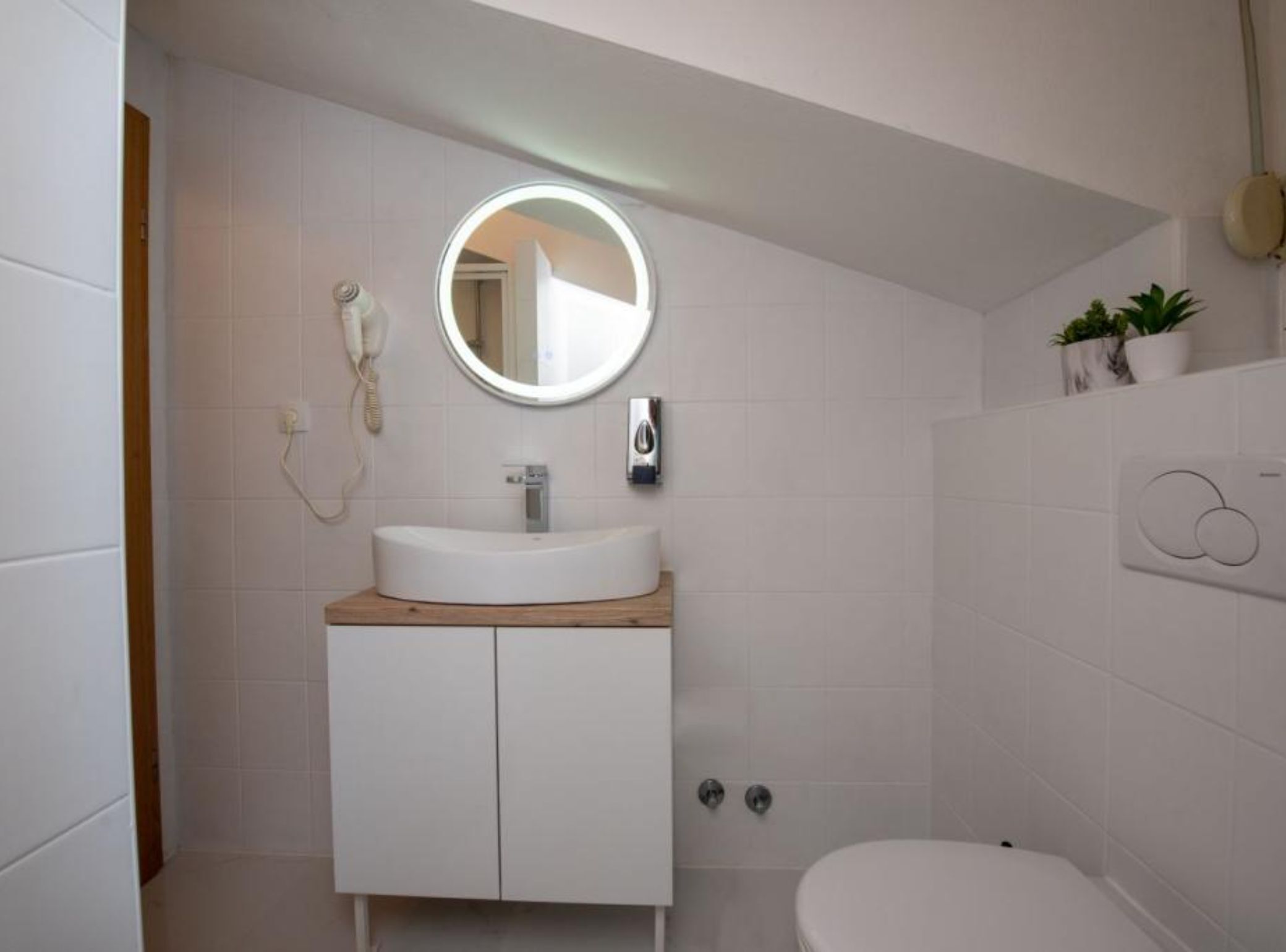 A modern bathroom with a round mirror, white tiles, a wooden countertop, a white sink, a toilet, and a wall-mounted heater.