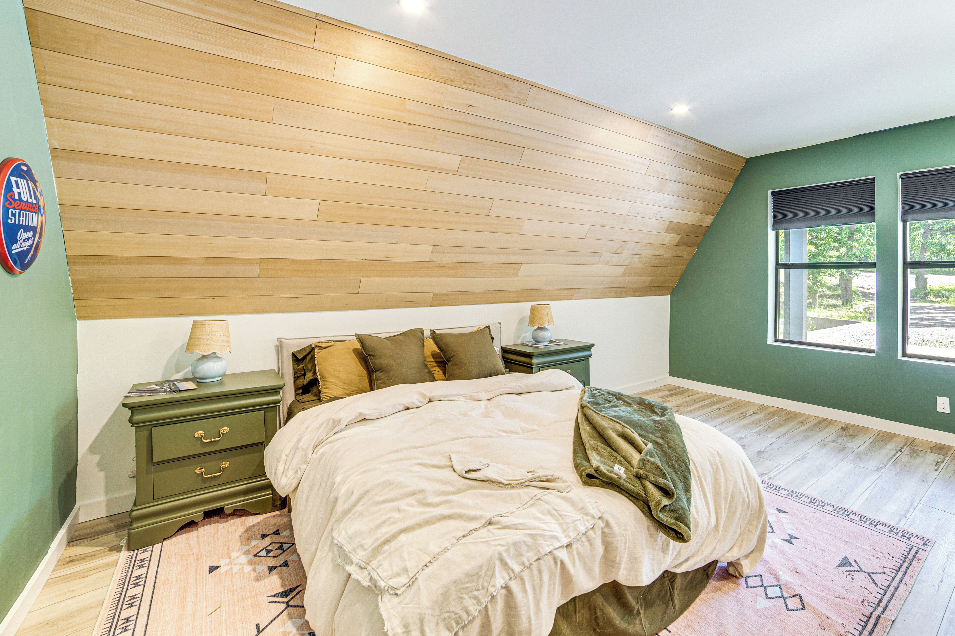 A bedroom with a wooden slat ceiling, a bed with a beige comforter, a nightstand with a lamp, and a view of the outdoors through a window.