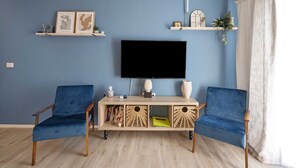 Un salon avec un mur bleu, un téléviseur à écran plat, deux fauteuils bleus, une table basse en bois et des étagères ornées d’objets décoratifs.