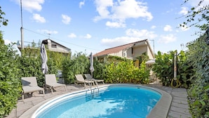 Une piscine entourée de chaises longues et de parasols, le tout bordé de verdure, avec une maison en arrière-plan.