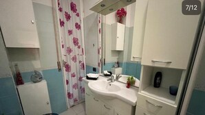 A bathroom with a white sink, mirror, and cabinets, a blue toilet, and a shower area with a floral curtain. - Charming 2-bedroom apartment with completed kitchen, nera Milano, Como and Swiss (Mozzate)