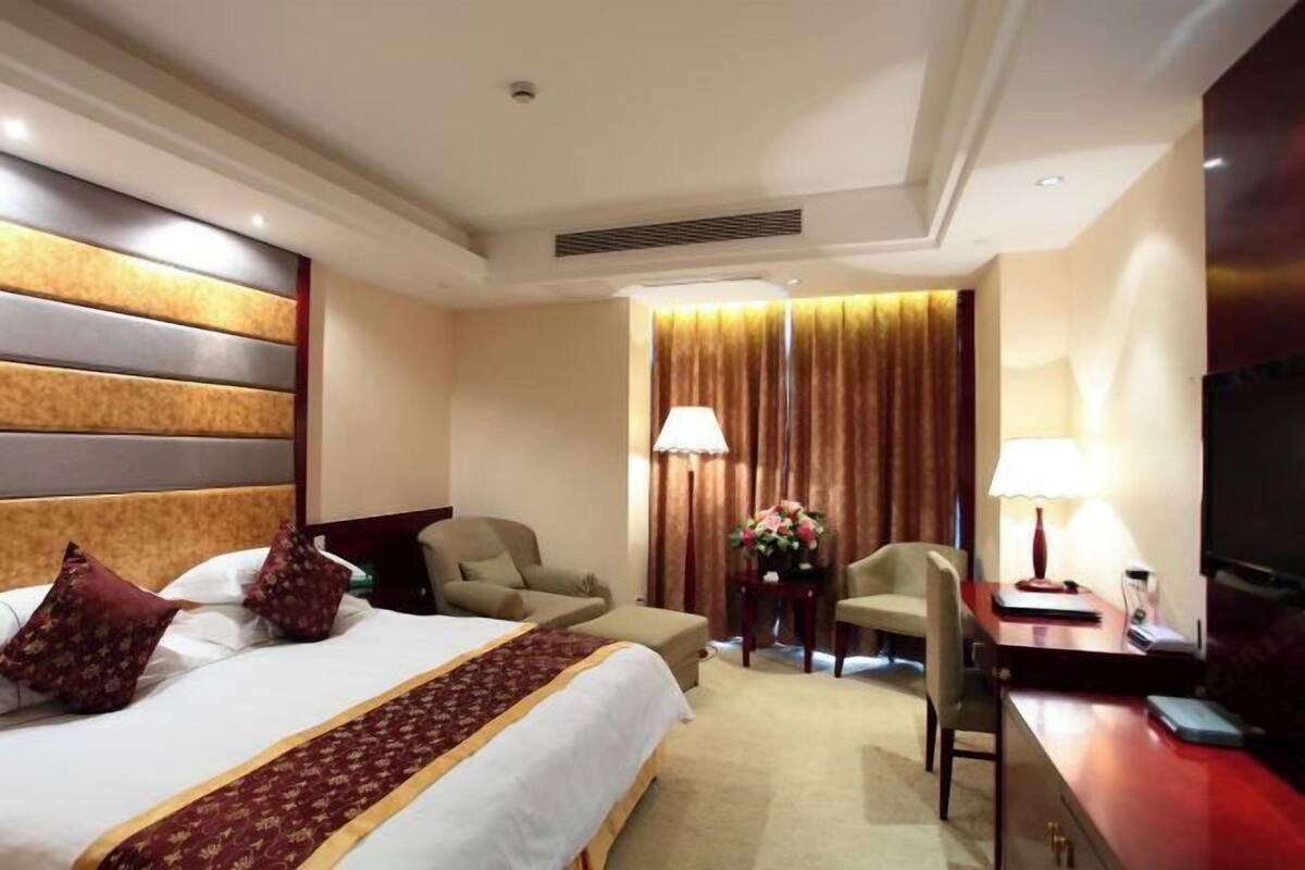 Une chambre d’hôtel avec un grand lit, deux chaises, un bureau, une télévision et une fenêtre avec des rideaux.