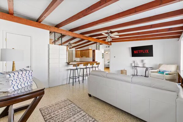A modern living room with a white sofa, a dining table with chairs, a kitchen area with wooden beams, and a wall-mounted Netflix sign.