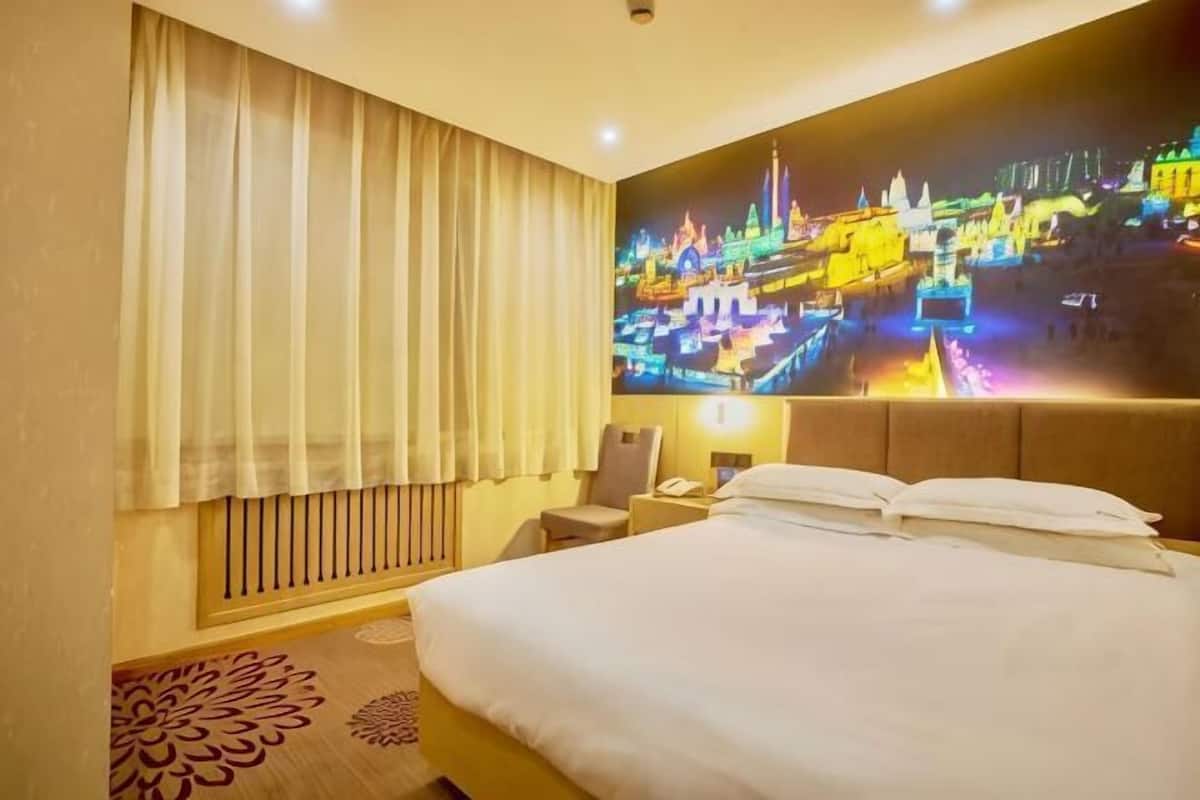 A hotel room with a large bed, a nightstand, a lamp, a window with curtains, and a wall-mounted TV displaying a cityscape.