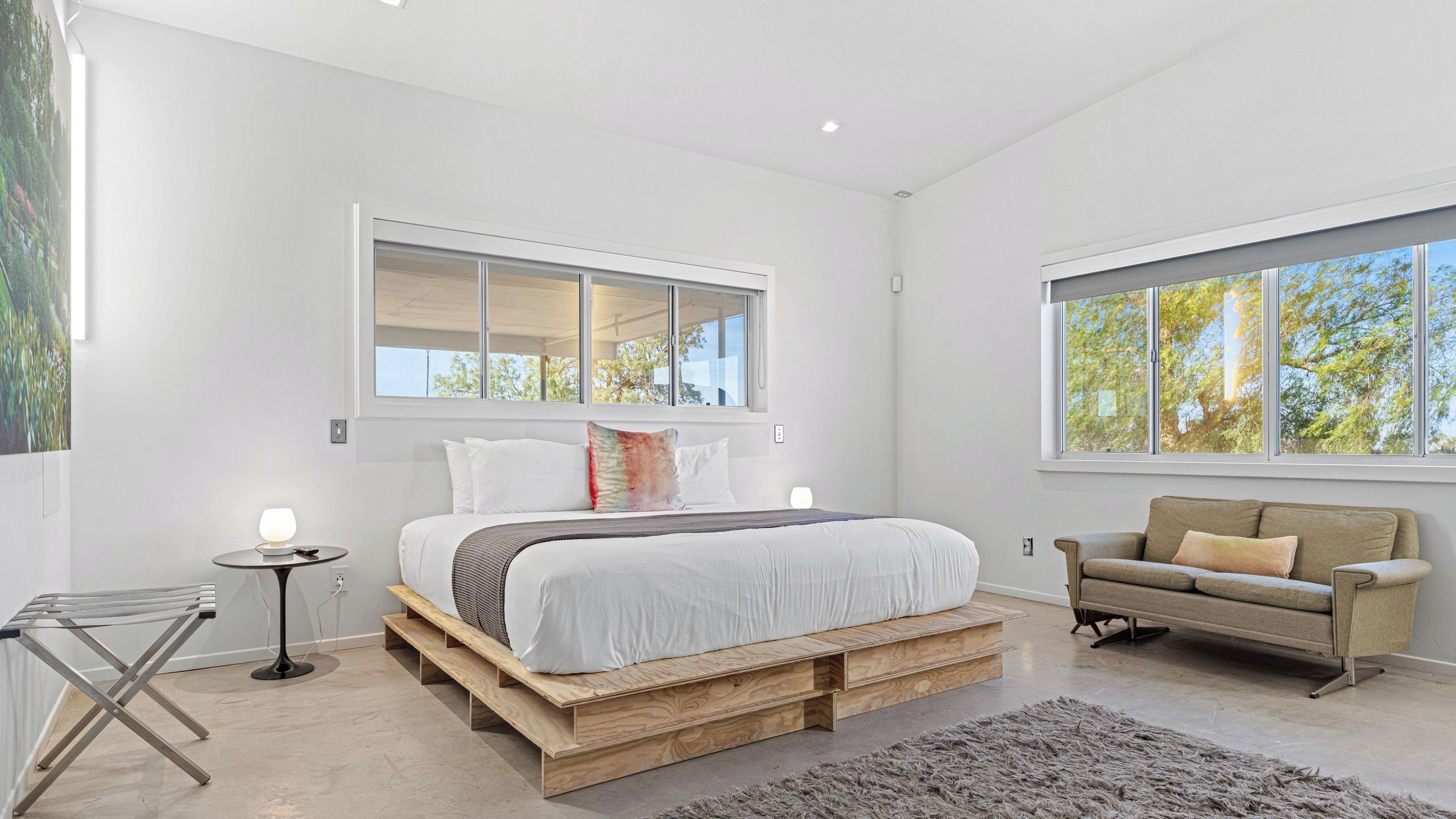 A modern bedroom with a bed on wooden pallets, a sofa, and large windows with natural light.