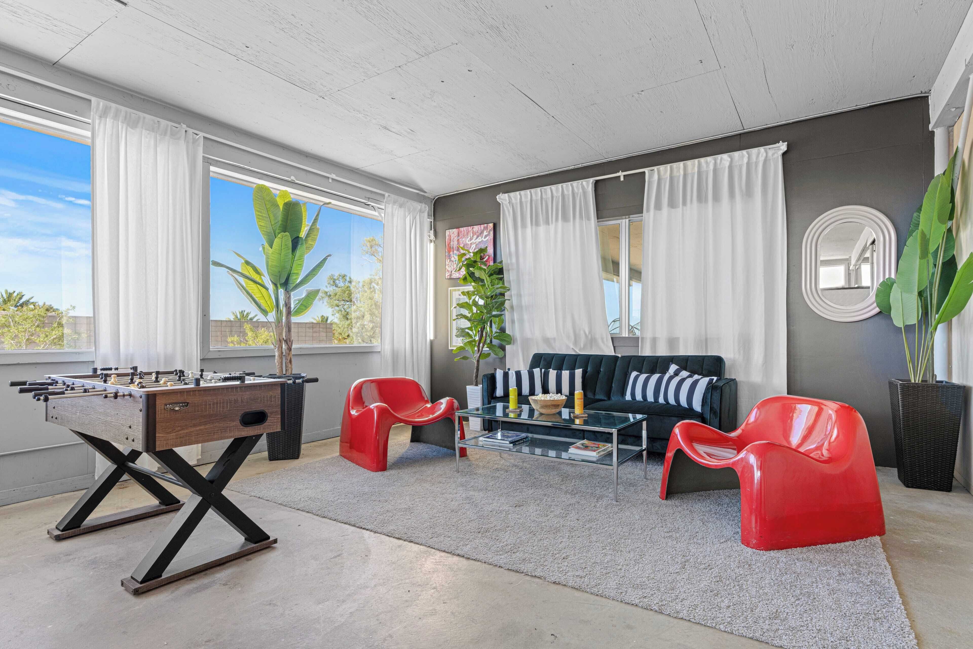 A modern living room with a foosball table, red chairs, a glass coffee table, and a sofa.