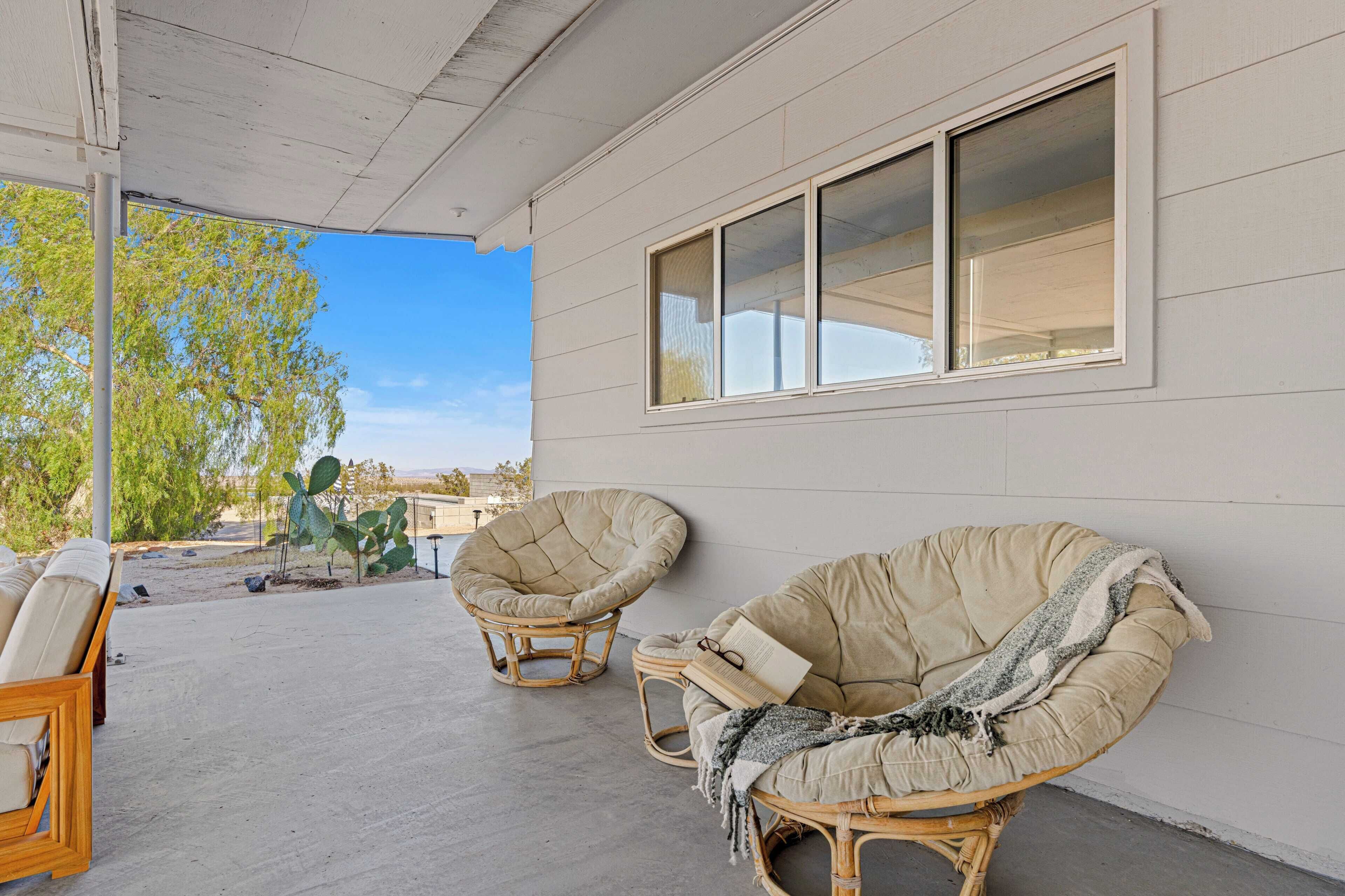 A covered patio area with two wicker chairs, one with a cushion and a blanket, and a wooden chair with cushions.
