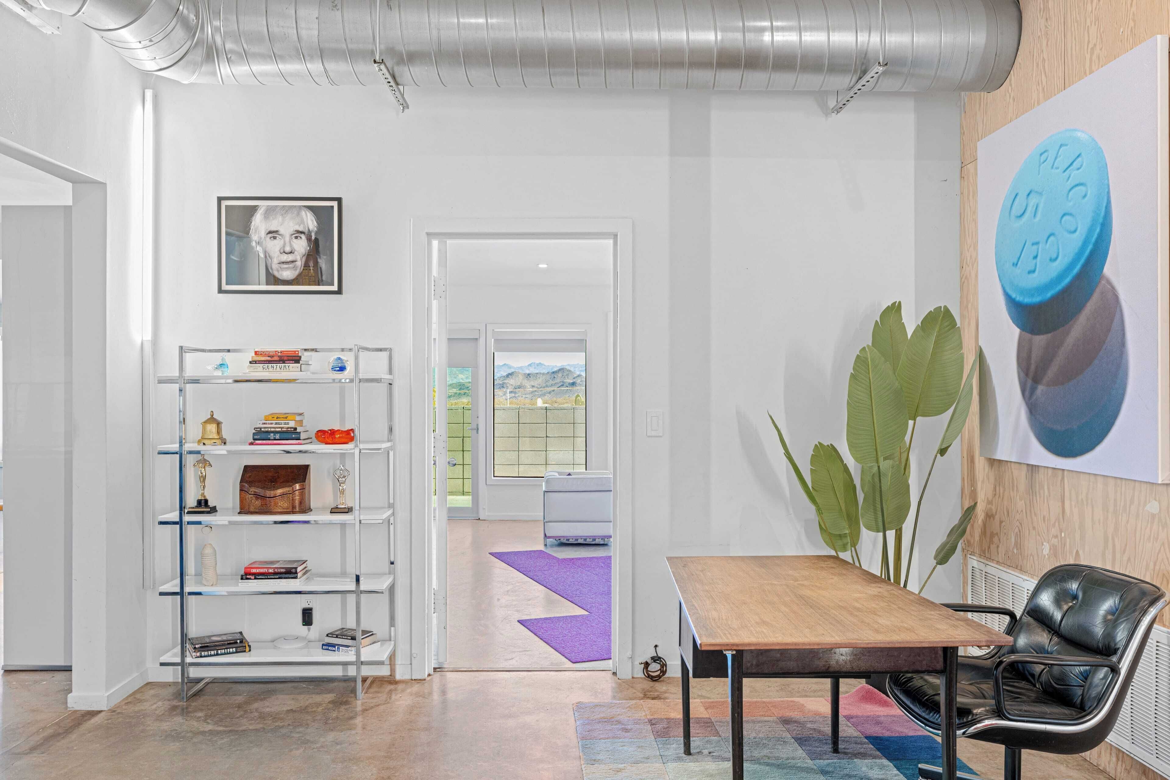 A modern interior with a wooden table and a leather chair, a bookshelf with various items, and a large plant.