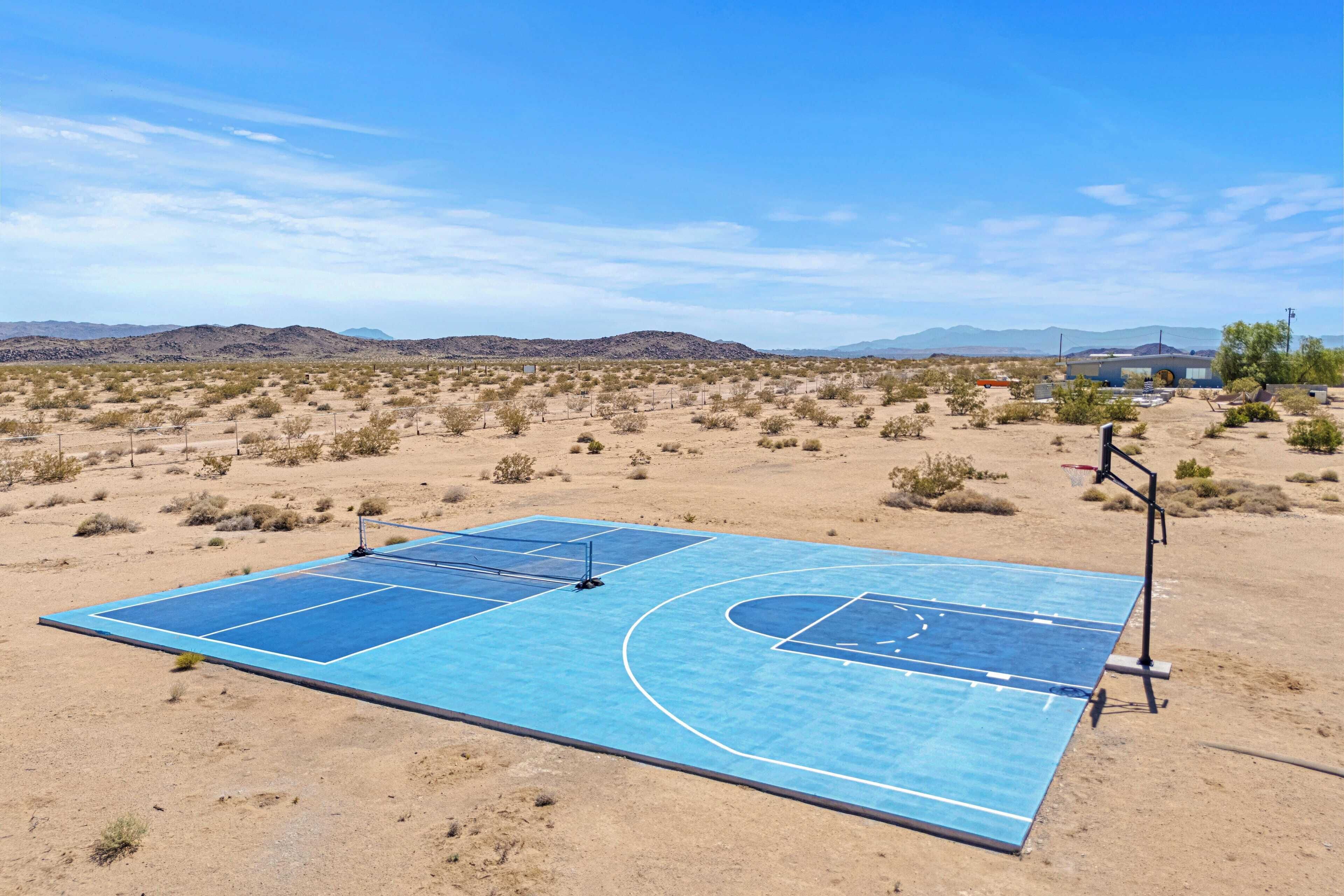 Un terrain de basket en plein air, doté d’une surface bleue, entouré de végétation désertique et avec des montagnes en arrière-plan.