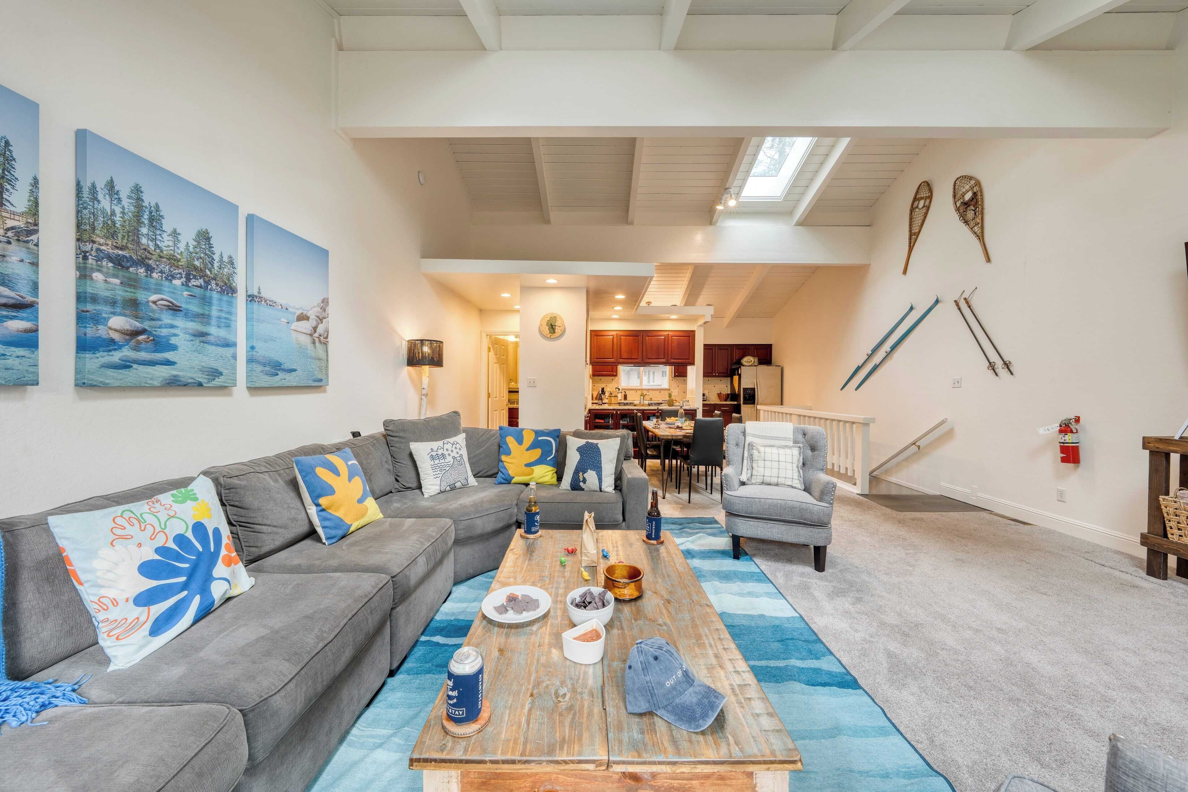 A spacious living room with a gray sectional sofa, a wooden coffee table with various items, and a blue rug. There are three paintings on the wall depicting a coastal scene. A kitchen and dining area are visible in the background.
