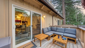 A cozy outdoor seating area with a sofa and two wooden tables, a glass door leading to a kitchen, and a wooden deck.