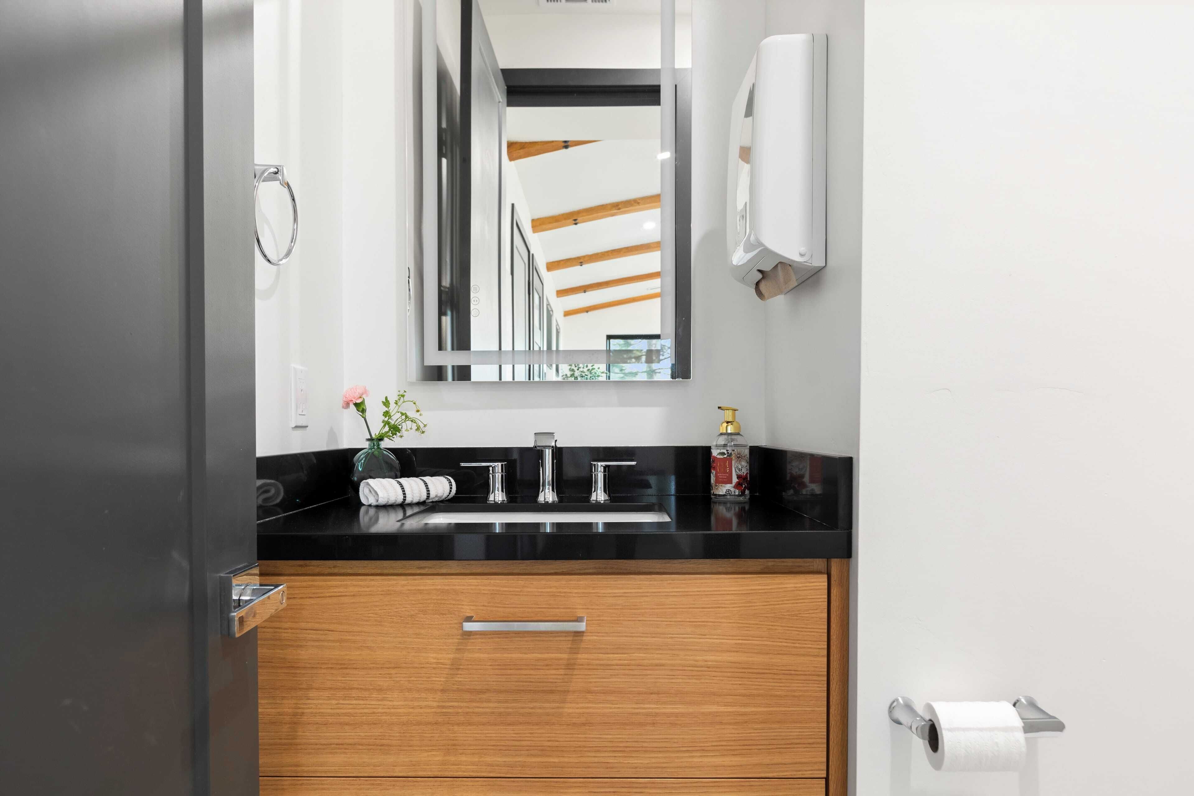 A modern bathroom with a black countertop, a wooden vanity, a mirror, a towel rack, a soap dispenser, and a toilet paper holder.